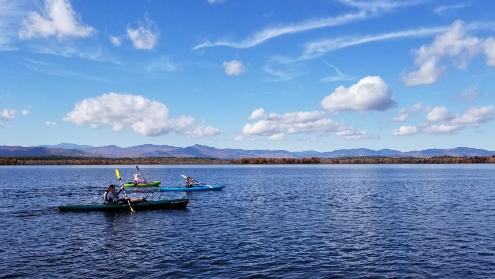 Kayakers paddle across lake with mountain and blue sky behind them